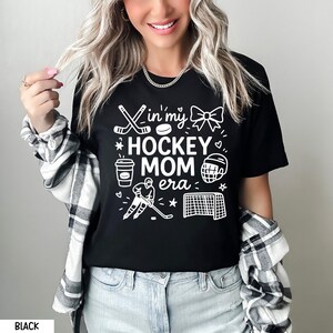 May include: Black t-shirt with white graphic text and illustrations. The text reads "In my Hockey Mom Era" with hockey-themed images. The shirt is worn with a plaid shirt and blue jeans.