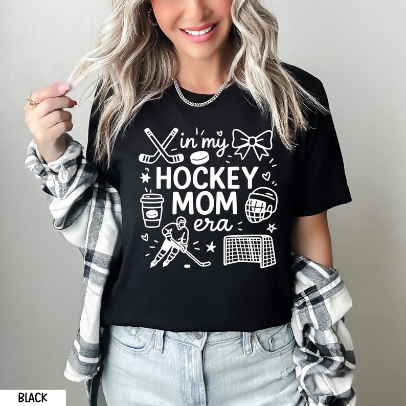 May include: Black t-shirt with white graphic text and illustrations. The text reads "In my Hockey Mom Era" with hockey-themed images. The shirt is worn with a plaid shirt and blue jeans.