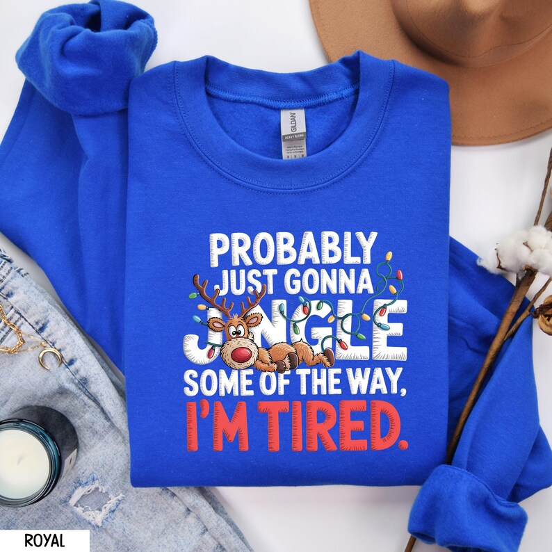 May include: Royal blue sweatshirt with the text "PROBABLY JUST GONNA JINGLE SOME OF THE WAY, I'M TIRED." A cartoon reindeer with antlers and a red nose is featured in the design, along with Christmas lights.