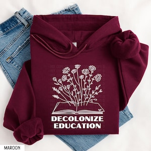 May include: A maroon hoodie with a white outline of an open book with flowers growing out of it. The text "DECOLONIZE EDUCATION" is printed below the book.