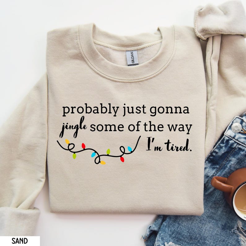 K&ouml;nnte beinhalten: Sandfarbenes Sweatshirt mit dem Text "probably just gonna jingle some of the way I'm tired." Der Text ist in schwarzer Schrift, darunter eine Lichterkette mit bunten Weihnachtslichtern.