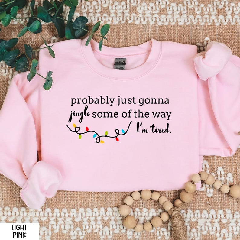 K&ouml;nnte beinhalten: Hellrosa Sweatshirt mit dem Text "probably just gonna jingle some of the way I'm tired." Der Text ist schwarz mit einer Lichterkette. Ein Gildan-Etikett ist sichtbar.