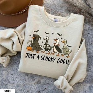 K&ouml;nnte beinhalten: Sandfarbenes Sweatshirt mit einem Halloween-Motiv. Das Design zeigt vier G&auml;nse in gruseligen Kost&uuml;men, mit Flederm&auml;usen und Sternen dar&uuml;ber. Der Text "JUST A SPOOKY GOOSE" ist darunter gedruckt.