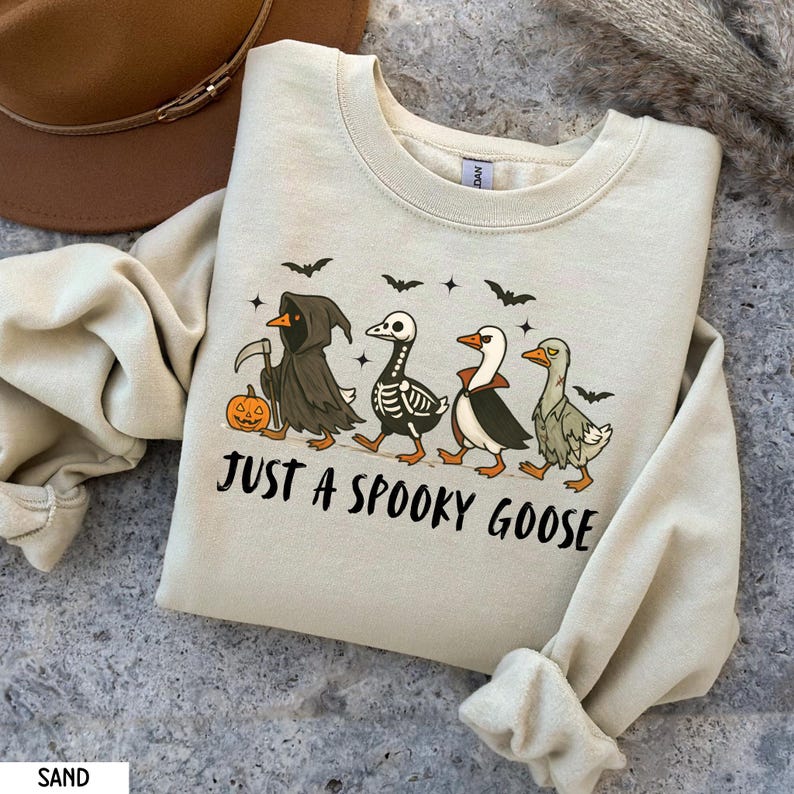 K&ouml;nnte beinhalten: Sandfarbenes Sweatshirt mit einem Halloween-Motiv. Das Design zeigt vier G&auml;nse in gruseligen Kost&uuml;men, mit Flederm&auml;usen und Sternen dar&uuml;ber. Der Text "JUST A SPOOKY GOOSE" ist darunter gedruckt.