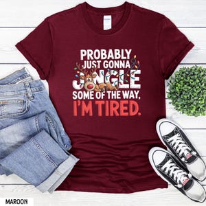 May include: A maroon t-shirt with the text "PROBABLY JUST GONNA JANGLE SOME OF THE WAY, I'M TIRED." The design includes a cartoon reindeer. The shirt is displayed with blue jeans and black and white trainers.