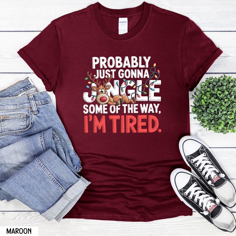 May include: A maroon t-shirt with the text "PROBABLY JUST GONNA JANGLE SOME OF THE WAY, I'M TIRED." The design includes a cartoon reindeer. The shirt is displayed with blue jeans and black and white trainers.