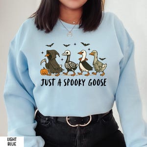 K&ouml;nnte beinhalten: Hellblaues Sweatshirt mit einem Halloween-Motiv. Das Design zeigt vier Cartoon-G&auml;nse in gruseligen Kost&uuml;men, darunter ein Sensenmann und ein Skelett. Der Text "JUST A SPOOKY GOOSE" steht unter den G&auml;nsen.
