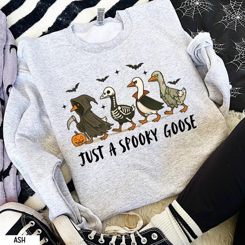 K&ouml;nnte beinhalten: Hellgraues Sweatshirt mit Halloween-Motiv. Das Design zeigt vier G&auml;nse in gruseligen Kost&uuml;men, Flederm&auml;use und den Text "JUST A SPOOKY GOOSE". Eine Gans ist als Sensenmann verkleidet und tr&auml;gt einen K&uuml;rbis.