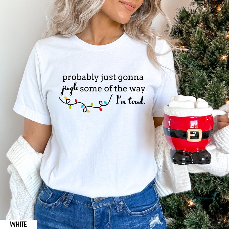 K&ouml;nnte beinhalten: Wei&szlig;es T-Shirt mit dem Text "probably just gonna jingle some of the way I'm tired." Der Text ist schwarz, mit dem Wort "jingle" in Schreibschrift und einer bunten Lichterkette. Eine Tasse im Santa-Design wird im Bild gehalten.
