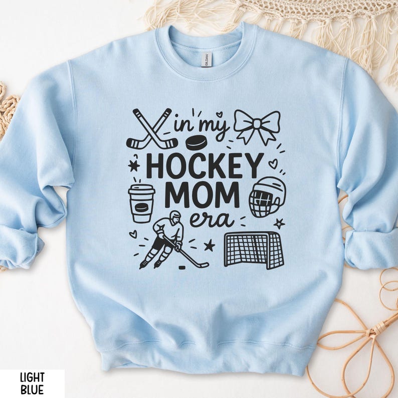 May include: Light blue sweatshirt with the text "In my Hockey Mom Era" and hockey-themed graphics. The design includes hockey sticks, a puck, a bow, a coffee cup, a hockey player, a helmet, and a net. The words "Light Blue" are at the bottom.