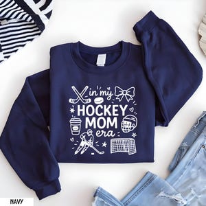 May include: Navy blue sweatshirt with the text "In my HOCKEY MOM era" in white. The design includes hockey sticks, a puck, a bow, a coffee cup, a hockey helmet, a hockey player, and a net. The sweatshirt is folded on a white surface.