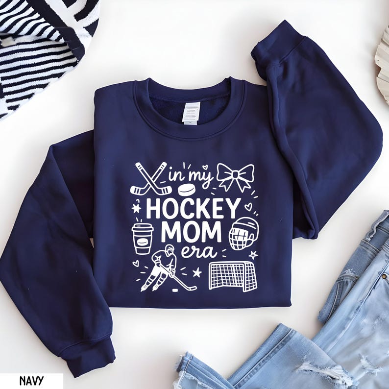 May include: Navy blue sweatshirt with the text "In my HOCKEY MOM era" in white. The design includes hockey sticks, a puck, a bow, a coffee cup, a hockey helmet, a hockey player, and a net. The sweatshirt is folded on a white surface.