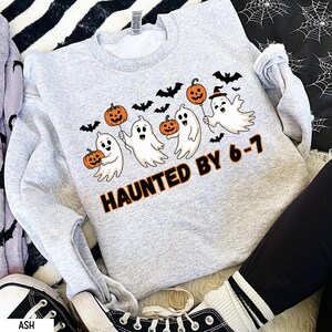 Peut inclure: Un sweat-shirt gris clair d'Halloween avec des fantômes de dessins animés tenant des ballons citrouilles oranges et des chauves-souris. Le texte "HAUNTED BY 6-7" est imprimé en orange et noir. Le sweat-shirt est de style col rond.