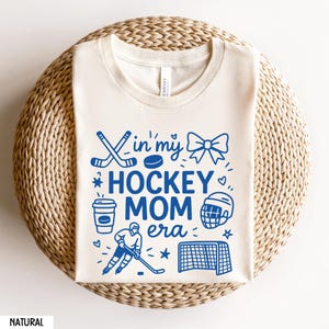 May include: Cream-colored t-shirt with the text "In my Hockey Mom Era" in blue, along with hockey-themed illustrations. The design includes hockey sticks, a puck, a bow, a coffee cup, a hockey player, a helmet, a net, and stars.
