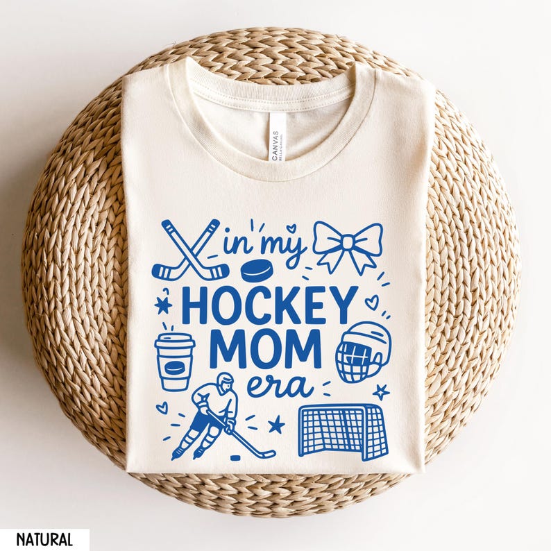 May include: Cream-colored t-shirt with the text "In my Hockey Mom Era" in blue, along with hockey-themed illustrations. The design includes hockey sticks, a puck, a bow, a coffee cup, a hockey player, a helmet, a net, and stars.