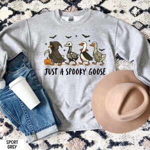 K&ouml;nnte beinhalten: Sportgraues Sweatshirt mit dem Text "JUST A SPOOKY GOOSE". Die Grafik zeigt vier Cartoon-G&auml;nse in Halloween-Kost&uuml;men, darunter ein Sensenmann, ein Skelett und ein Vampir. Ein K&uuml;rbis und Flederm&auml;use sind ebenfalls im Design enthalten.