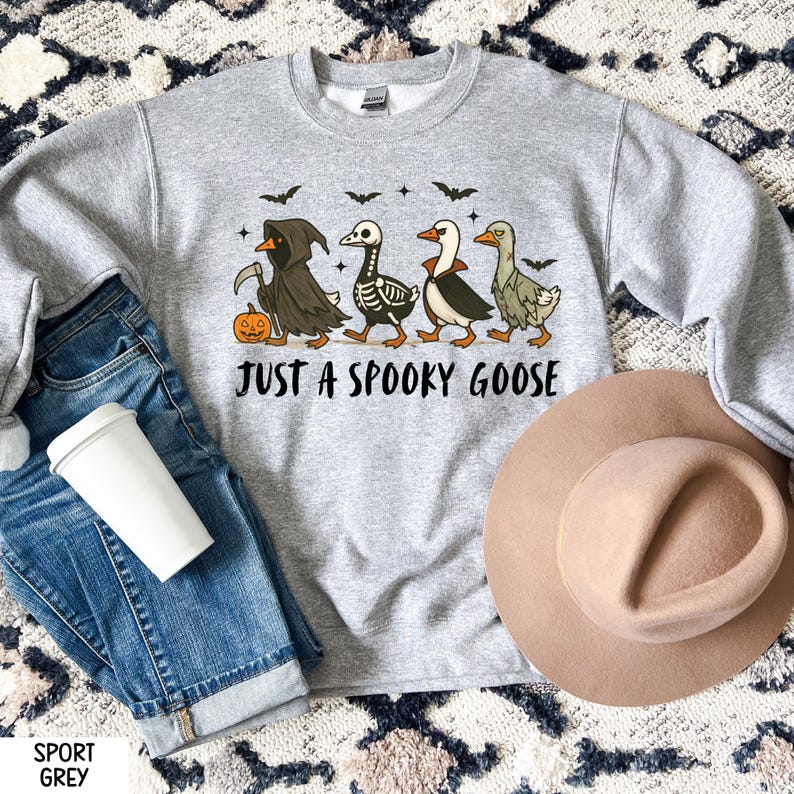 K&ouml;nnte beinhalten: Sportgraues Sweatshirt mit dem Text "JUST A SPOOKY GOOSE". Die Grafik zeigt vier Cartoon-G&auml;nse in Halloween-Kost&uuml;men, darunter ein Sensenmann, ein Skelett und ein Vampir. Ein K&uuml;rbis und Flederm&auml;use sind ebenfalls im Design enthalten.