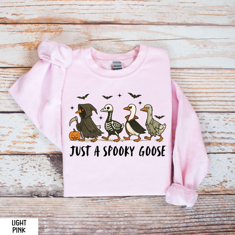K&ouml;nnte beinhalten: Hellrosa Sweatshirt mit einem Halloween-Motiv. Das Design zeigt vier Cartoon-G&auml;nse in gruseligen Kost&uuml;men, darunter ein Sensenmann, ein Skelett und ein Vampir. Der Text "JUST A SPOOKY GOOSE" ist darunter gedruckt.