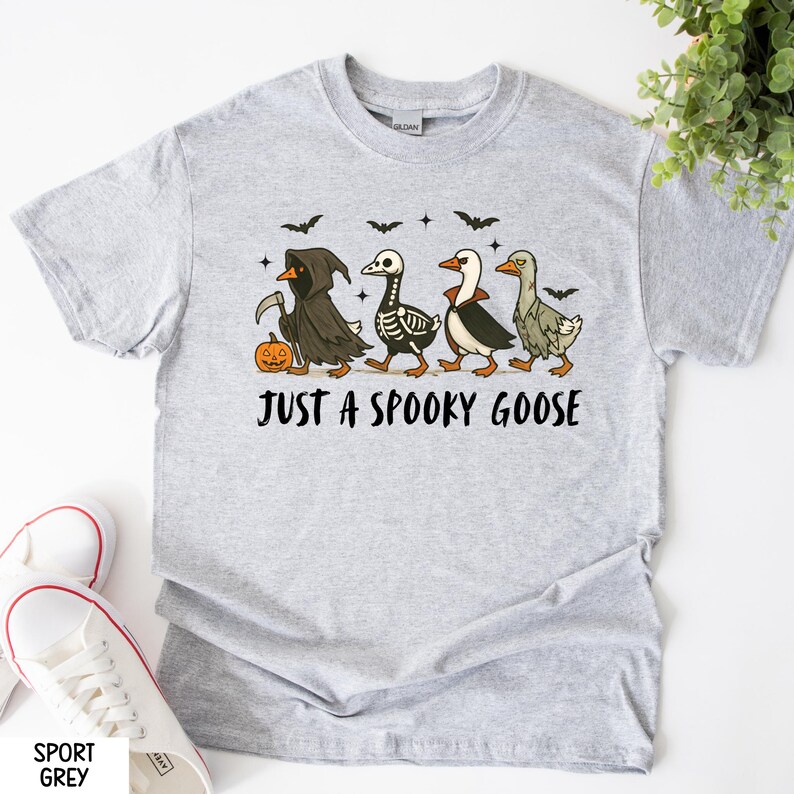 K&ouml;nnte beinhalten: Hellgraues T-Shirt mit einem Halloween-Motiv. Das Design zeigt vier G&auml;nse in gruseligen Kost&uuml;men, darunter ein Sensenmann, ein Skelett und ein Vampir. Der Text "JUST A SPOOKY GOOSE" ist darunter gedruckt.