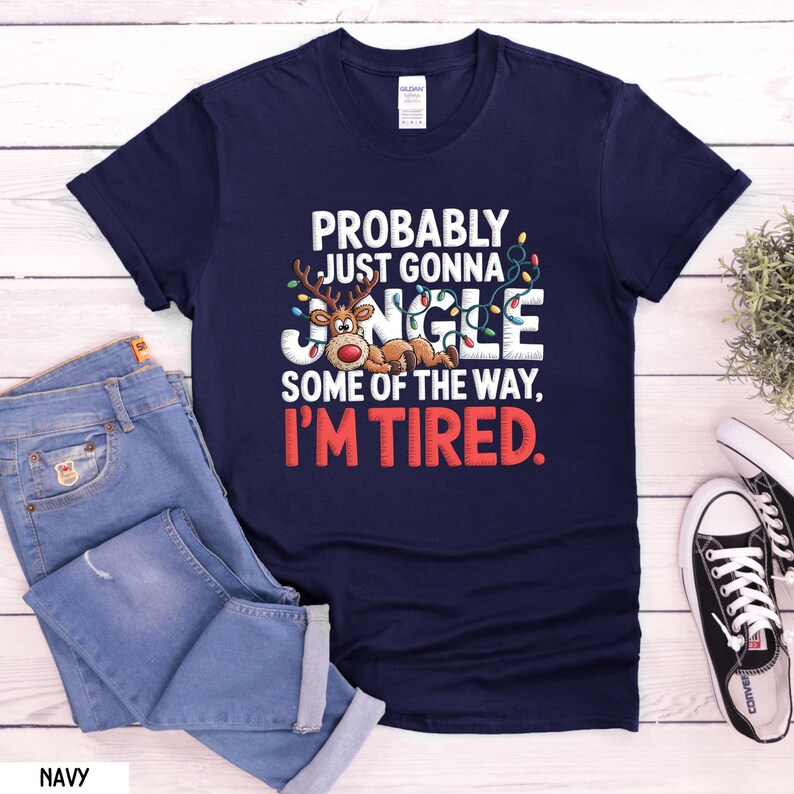 May include: Navy blue t-shirt with the text "PROBABLY JUST GONNA JINGLE SOME OF THE WAY, I'M TIRED." A cartoon reindeer is featured in the design, along with Christmas lights. The shirt is paired with blue jeans and white trainers.