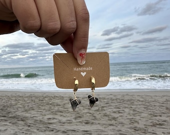 May include: Gold hoop earrings with dark gray shark teeth pendants, displayed on a brown card that reads "Handmade". The earrings are held up against a beach backdrop with a cloudy sky and ocean waves.