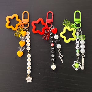 May include: Three handmade keychains with star-shaped clasps in orange, red, and green. Each keychain features a unique design with beads, charms, and lettering spelling out "ATEEZ", "SKZ STAY", and "NCT".