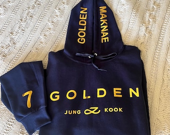 May include: Navy blue hoodie with a gold embroidered logo that says "Golden Jung Kook" and the number "7" on the sleeve. The hood has the words "Golden" and "Maknae" embroidered on the sides.