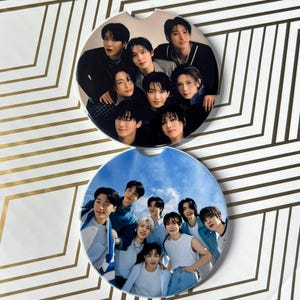 May include: Two round, white ceramic coasters with images of a K-pop boy band. The top coaster features the band members in a studio setting, while the bottom coaster shows them outdoors against a blue sky.