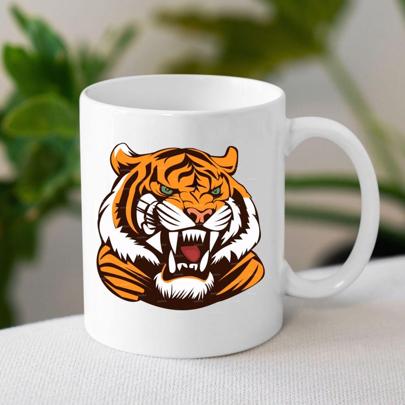 May include: White ceramic coffee mug with a graphic of a tiger's face with orange and black stripes and a white background. The tiger has a fierce expression with its mouth open and teeth showing.