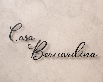 May include: Black metal sign with the words "Casa Bernardina" in elegant cursive script. The sign is mounted on a textured, light beige wall, creating a contrast between the dark lettering and the neutral background.