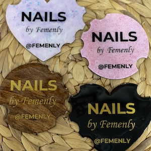 May include: Four heart-shaped nail art decals with the text "NAILS by Femenly @FEMENLY" in gold lettering. The decals are in various colors, including pink, purple, brown, and black.