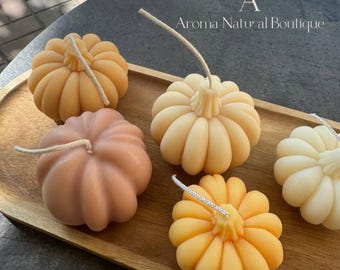 May include: Four small pumpkin-shaped candles on a wooden tray. The candles are in shades of orange, yellow, and brown. The text "Aroma Natural Boutique" is visible in the top right corner of the image.