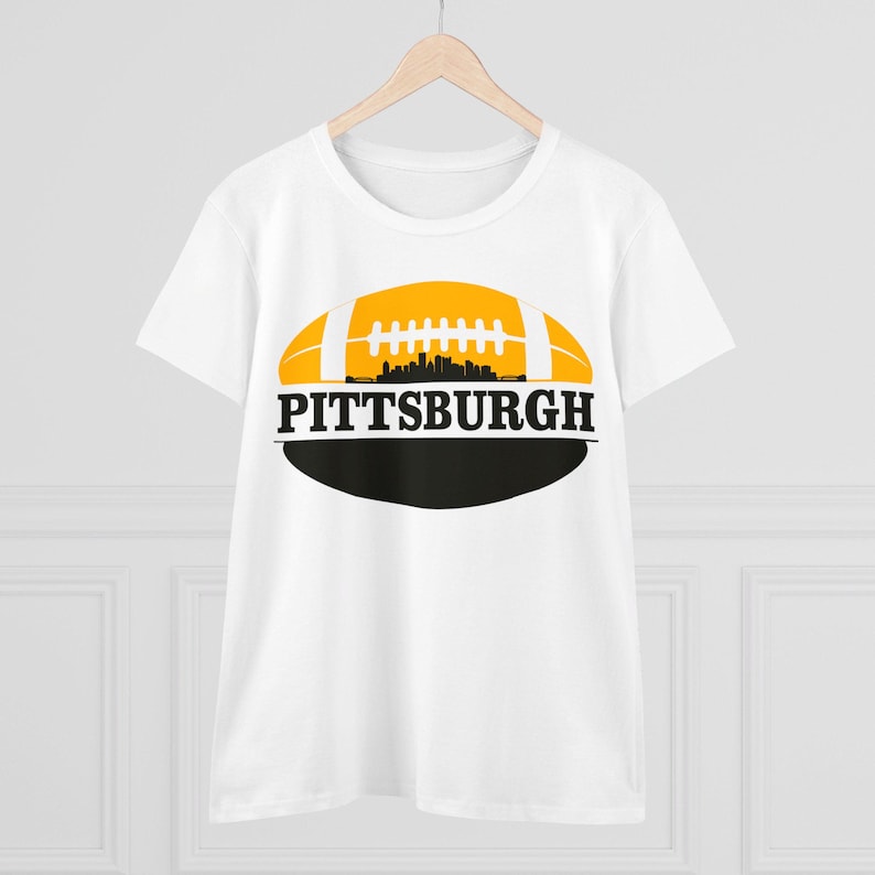 May include: White t-shirt with a yellow and black football design. The football has a silhouette of the Pittsburgh skyline inside. The text "PITTSBURGH" is printed below the football.