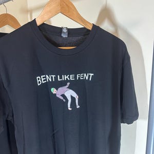 Puede incluir: Camiseta negra con un gráfico blanco que dice "Bent Like Fent" y una ilustración de dibujos animados de una persona inclinándose.