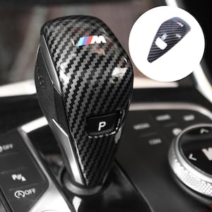 May include: A gear shift knob cover featuring a black and gray carbon fiber pattern with a glossy finish. The cover displays the M logo with blue, purple, and red stripes and a P button. A circular inset shows the cover isolated on a white background.