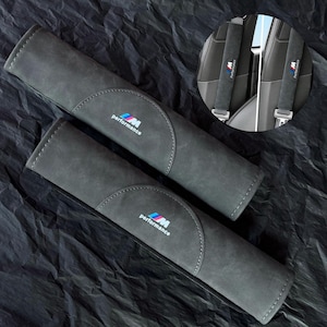 Puede incluir: Dos fundas grises para cinturón de seguridad con el logotipo de BMW M Performance en azul, rojo y blanco. Las fundas están hechas de un material suave y tienen un diseño cosido. La imagen también muestra un primer plano del cinturón de seguridad con el mismo logotipo.
