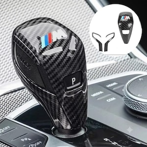 May include: A carbon fiber gear shift knob cover featuring the M logo in blue, red, and white. The cover has a glossy finish, the word UNLOCK embossed on the side, and a P button. An inset shows the cover and two trim pieces from a top view.