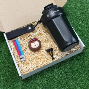 Könnte beinhalten: Ein schwarzer Shaker mit M Power-Logo, eine schwarze Stoffhandschlaufe, ein Schlüsselanhänger mit roten, blauen und hellblauen Streifen und M-Logo, ein brauner bärenförmiger Anhänger mit blau-roter Kappe und ein kleiner schwarzer Clip.