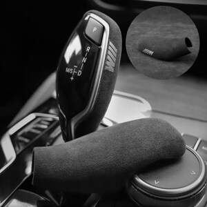以下が含まれることがあります： Mロゴが入ったダークグレーのスエード調シフトノブカバー。車両のギアセレクターにフィットするデザインで、柔らかい質感の表面が特徴です。P、R、N、Dの表示があるシフトレバーに装着された状態と、単体の状態の両方が示されています。