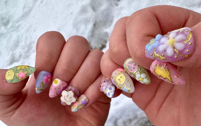 May include: Close-up of a hand with colourful, decorated fingernails. The nails are painted in various pastel colours and feature designs such as flowers, smiley faces, stars, and peace signs.