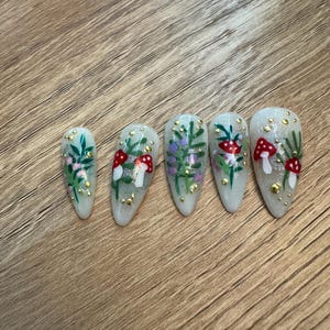 May include: Five white oval-shaped false nails with a woodland theme. Each nail has a different design featuring green leaves, red and white mushrooms, and gold glitter.