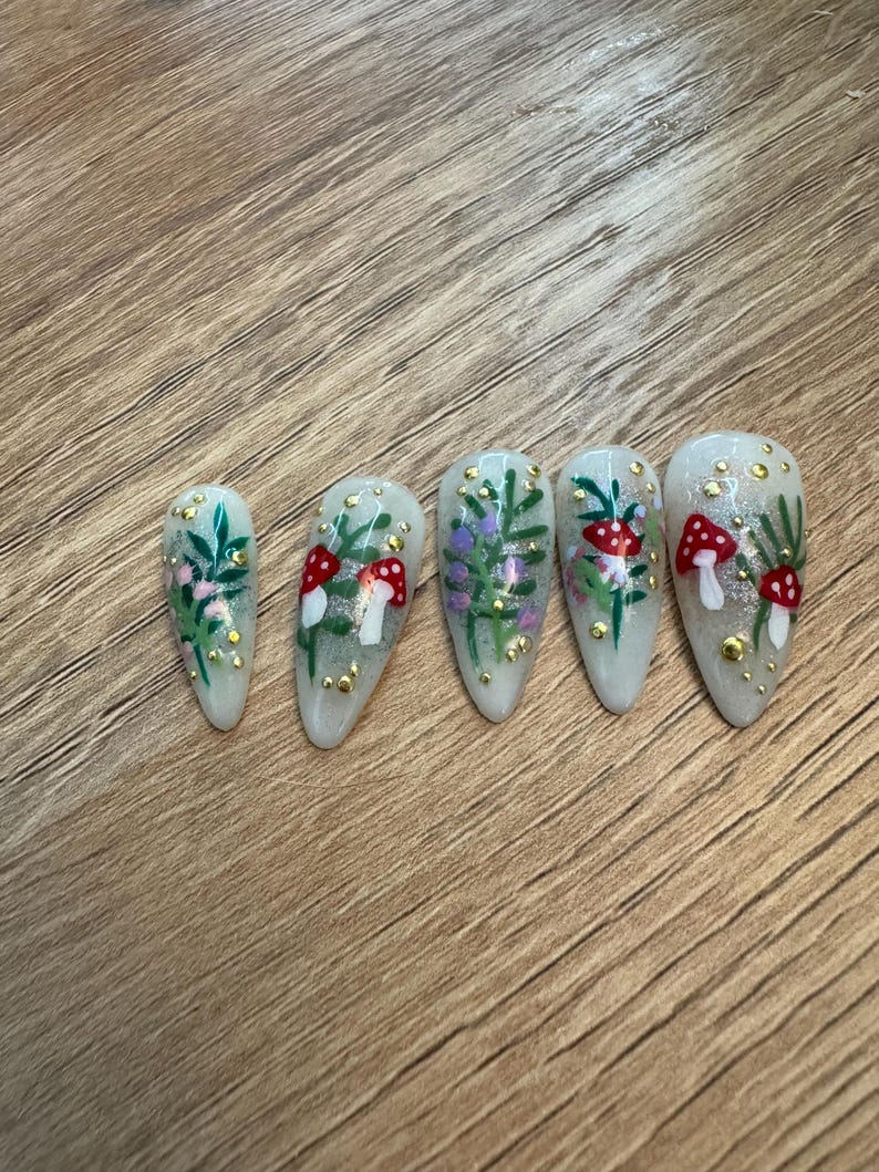May include: Five white oval-shaped false nails with a woodland theme. Each nail has a different design featuring green leaves, red and white mushrooms, and gold glitter.