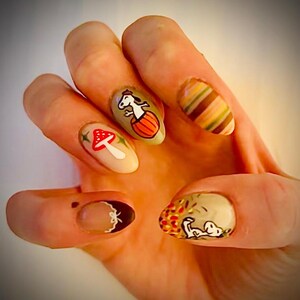 May include: Close-up of a hand with autumn-themed nail art. The nails are painted in various shades of brown, beige, and white, with designs of mushrooms, a pumpkin, and a striped pattern.