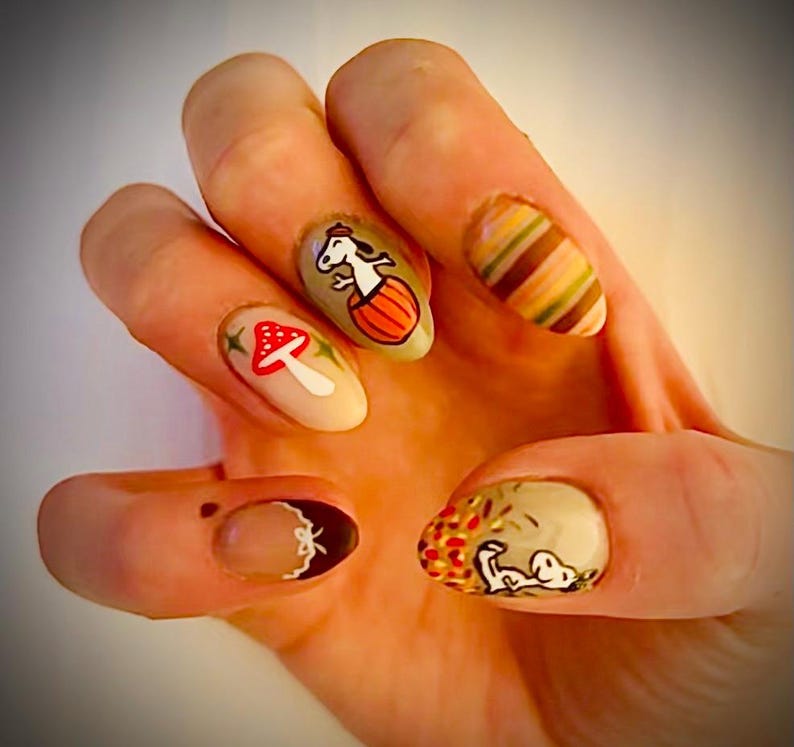 May include: Close-up of a hand with autumn-themed nail art. The nails are painted in various shades of brown, beige, and white, with designs of mushrooms, a pumpkin, and a striped pattern.
