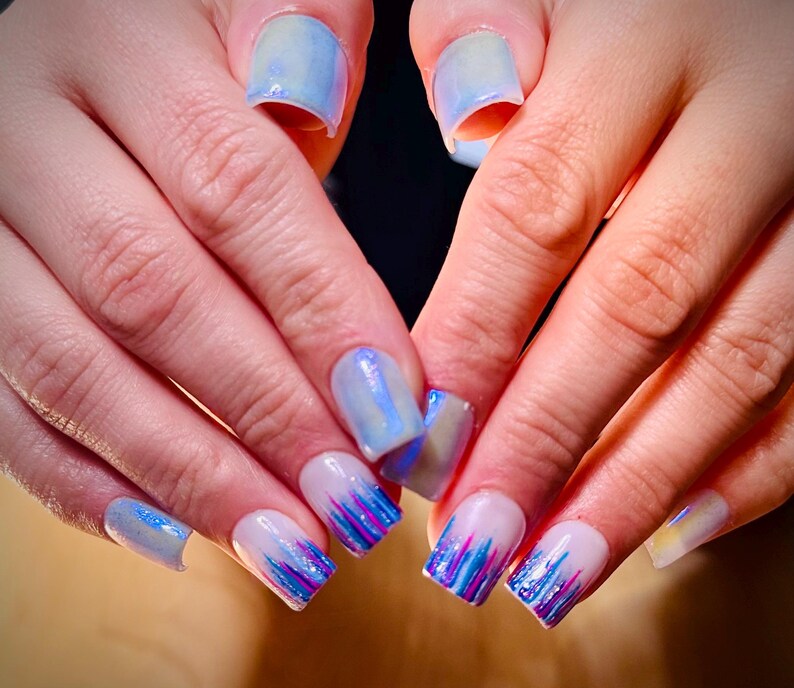 May include: Close-up of a hand with blue and white nail polish. The nails are decorated with a blue and white marble design and a blue and purple glitter design.