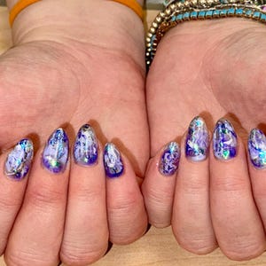 May include: Close-up of hands with purple and silver nail polish. The nails are long and almond shaped, with a marbled design.