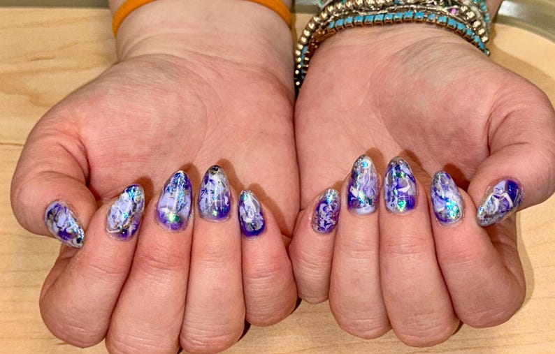 May include: Close-up of hands with purple and silver nail polish. The nails are long and almond shaped, with a marbled design.