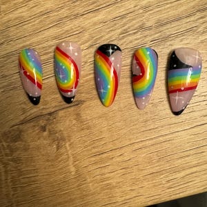 May include: Five false nails painted with a rainbow design, featuring white stars and black accents. The nails are displayed in a row on a wooden surface.