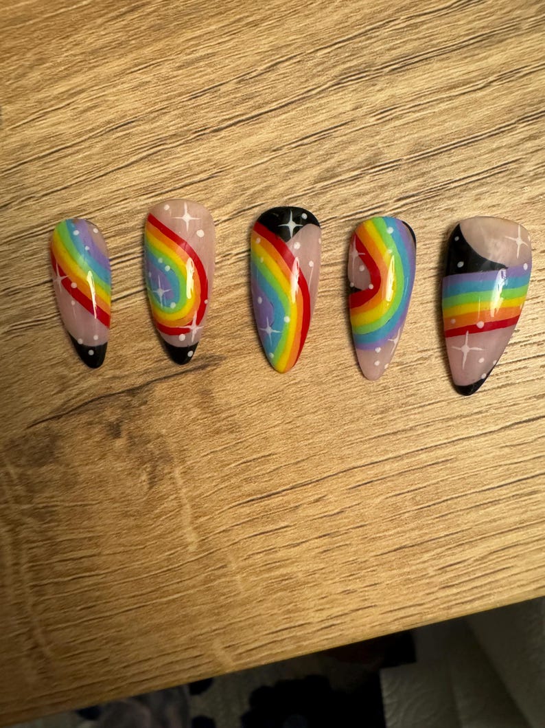 May include: Five false nails painted with a rainbow design, featuring white stars and black accents. The nails are displayed in a row on a wooden surface.