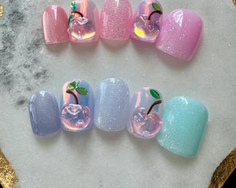 May include: A collection of ten artificial nails in shades of pink, blue, and purple. Some nails are glittery, while others have a 3D cherry design with green leaves and a brown stem. The nails are displayed on a white surface.